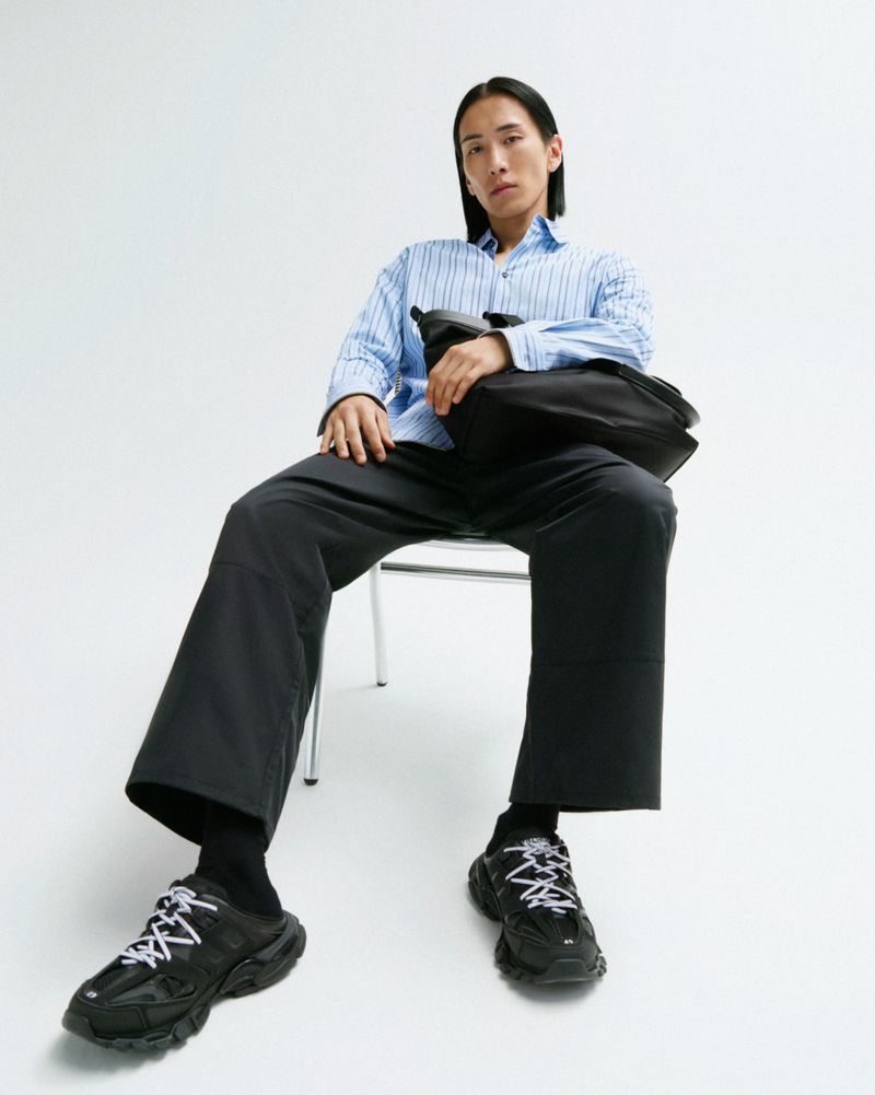 An adult male sits on a chair in a photo studio, wearing a blue striped shirt, black trousers, black socks, and black sneakers.