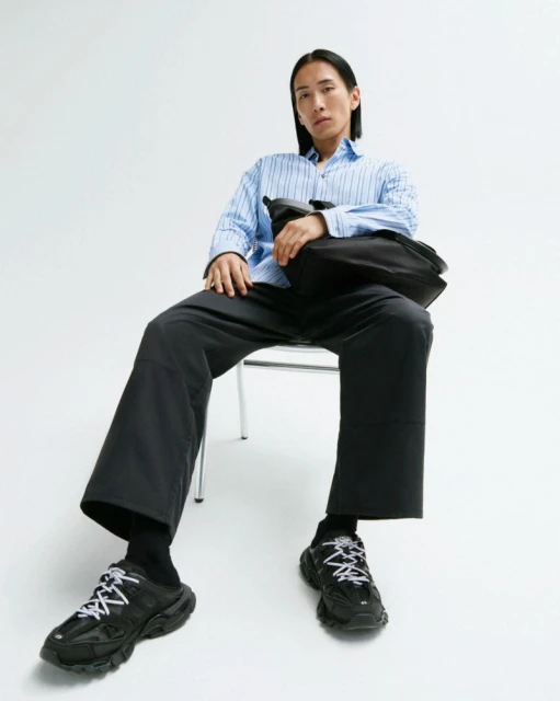 An adult male sits on a chair in a photo studio, wearing a blue striped shirt, black trousers, black socks, and black sneakers.