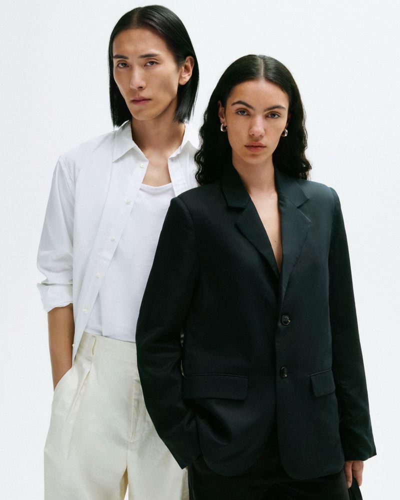 Two fashion models pose in front of a white backdrop. The model on the right wears a black blazer, while the other is in white.