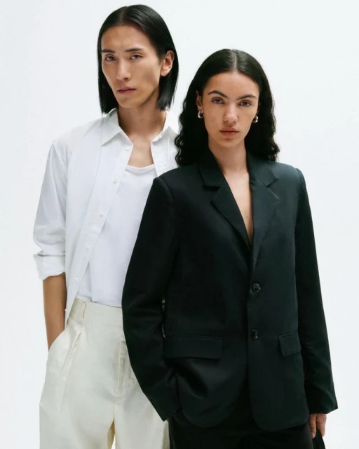 Two fashion models pose in front of a white backdrop. The model on the right wears a black blazer, while the other is in white.