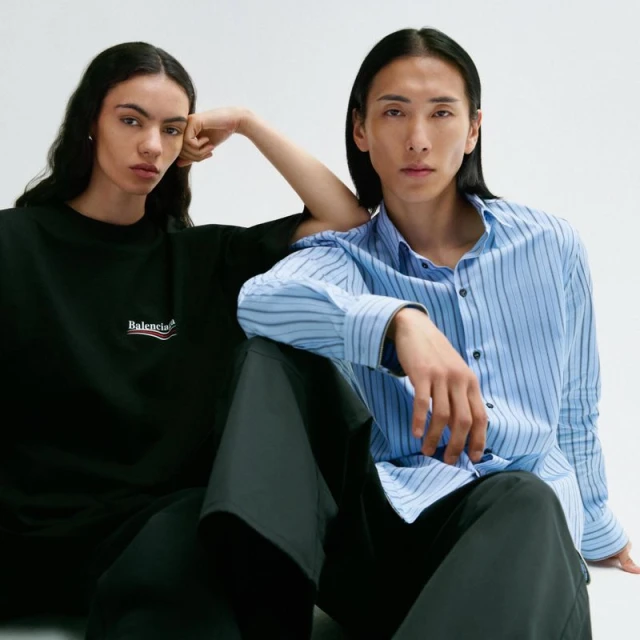 Two fashion models, one woman and one man, pose in front of a white background, wearing fashionable clothing. The woman wears black, the man wears blue.
