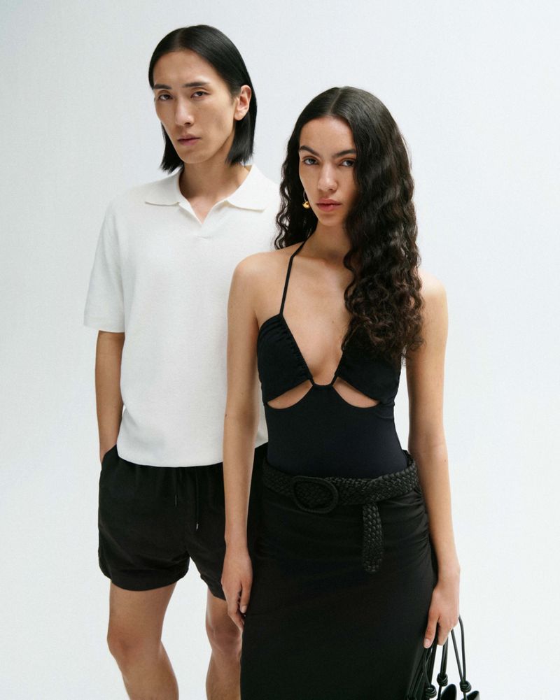 Two fashion models pose in front of a bright white backdrop, the woman wears a black bodysuit with a skirt and the man wears a white polo shirt.