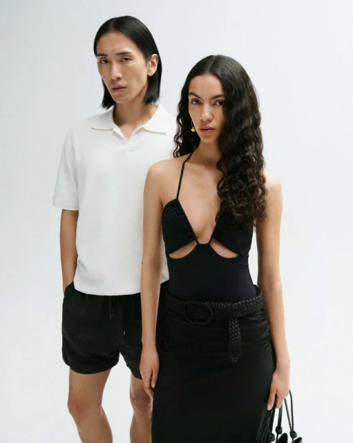 Two fashion models pose in front of a bright white backdrop, the woman wears a black bodysuit with a skirt and the man wears a white polo shirt.