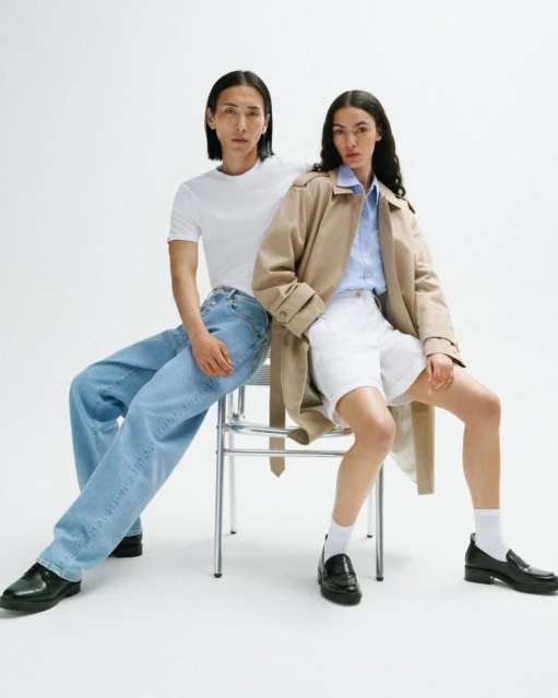 Two models sit on a chair in a studio setting. One wears a white t-shirt and jeans, the other a trench coat, shorts, and a blue shirt.