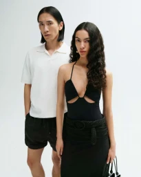 A man in a white polo and black shorts standing beside a woman in a black halter top and skirt against a seamless white background.