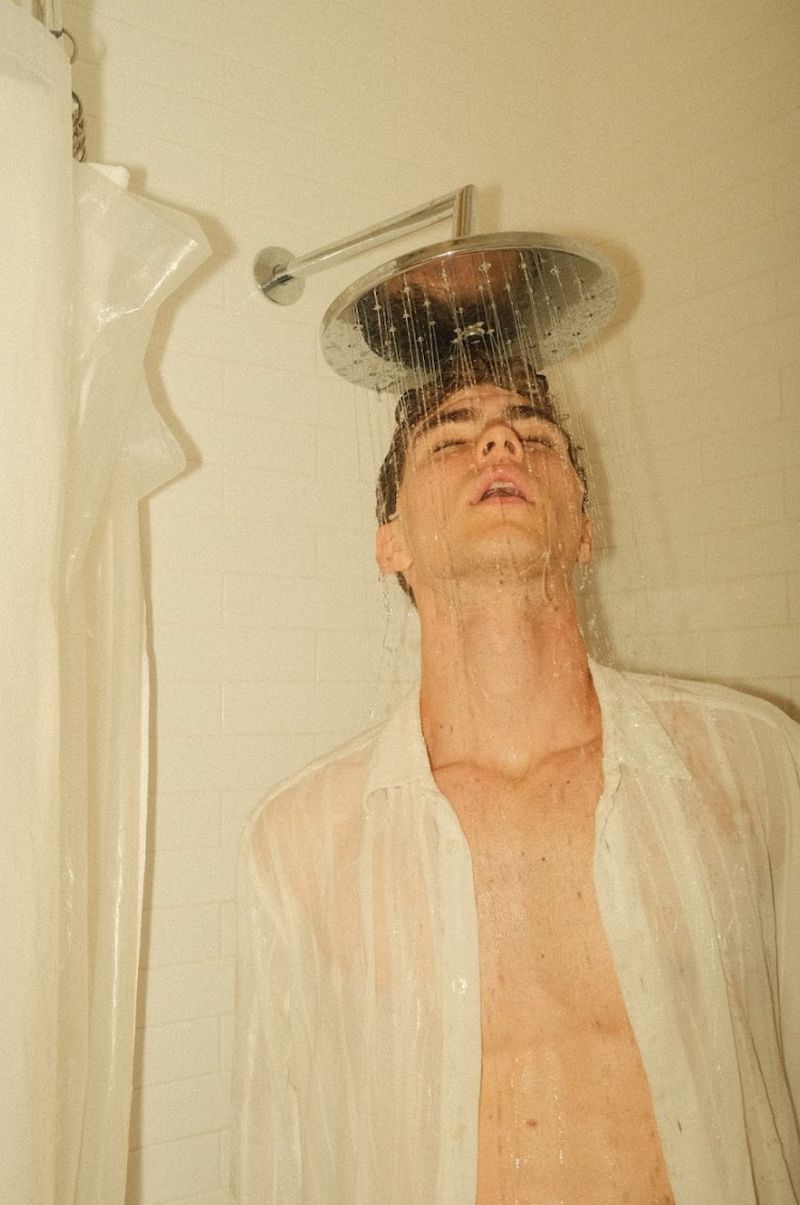 Lucas Tarrago standing under a streaming showerhead wearing a wet unbuttoned white shirt against tiled bathroom walls.