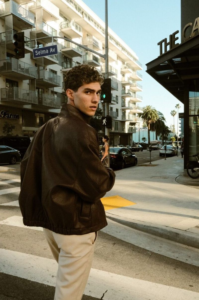 Lucas Tarrago wearing an oversized brown leather jacket and cream trousers walks across a city crosswalk, glancing back.