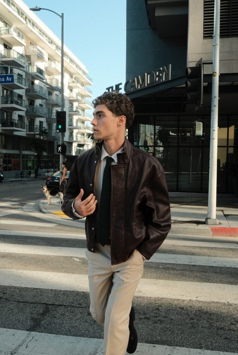 Lucas Tarrago walks across an urban crosswalk wearing a dark brown leather jacket, collared shirt with a black tie, and beige trousers.