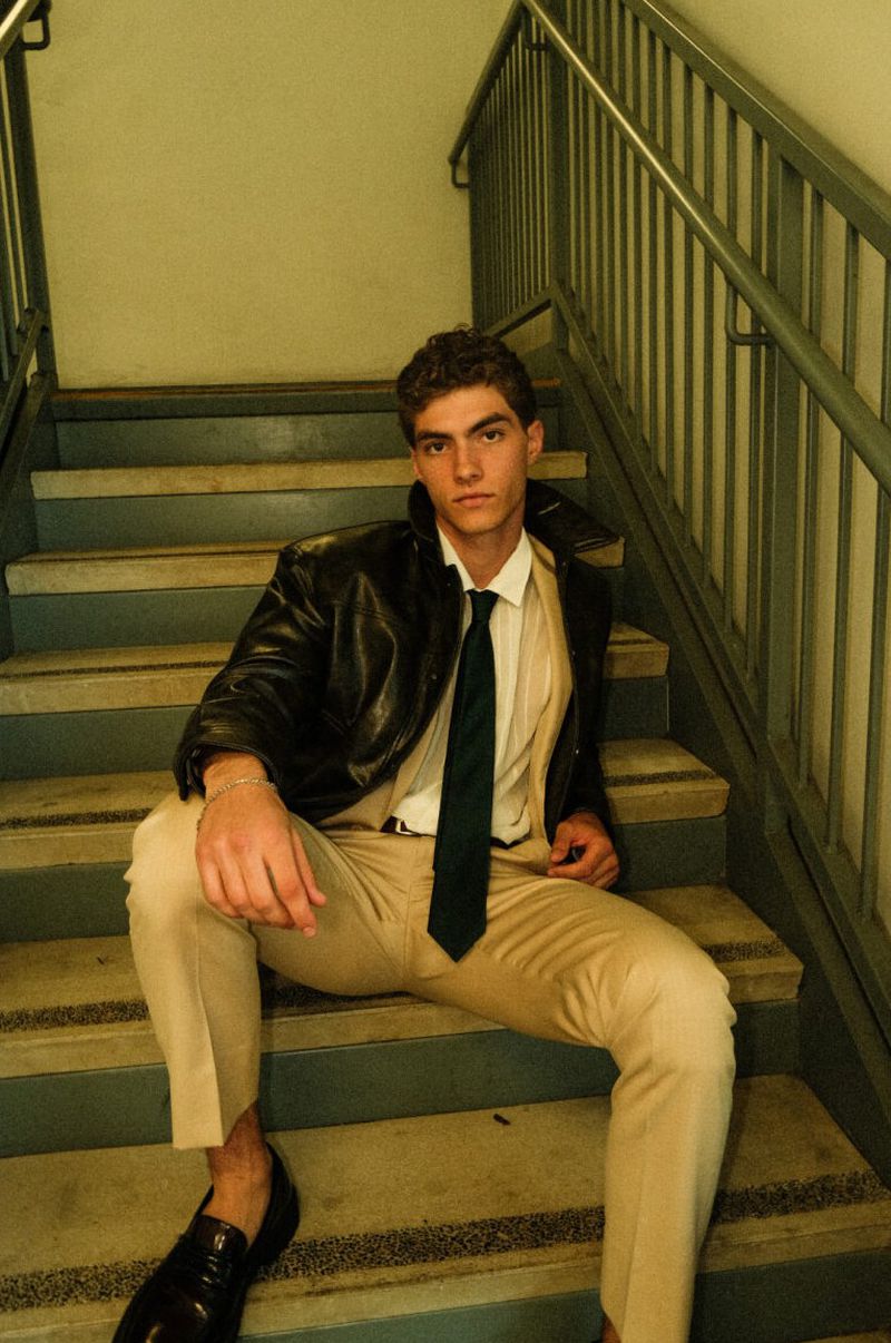 Model Lucas Tarrago sitting on an indoor staircase wearing a black leather jacket, beige trousers, and a tie for Vanity Teen Magazine.