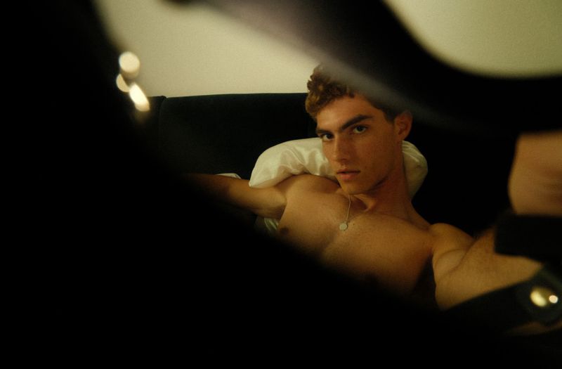 Lucas Tarrago reclining shirtless against a white pillow and dark headboard in an intimate, low-key portrait.