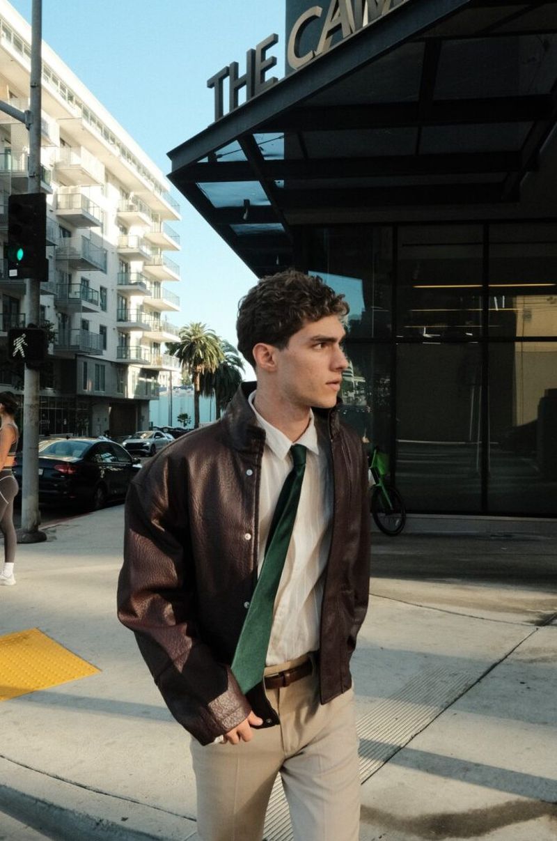 Model Lucas Tarrago walking along a city sidewalk wearing an oversized brown leather jacket, green tie, and beige trousers.