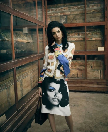 Model Nora Attal posing in a printed jacket and graphic face-print skirt inside a museum with tall wooden glass vitrines.