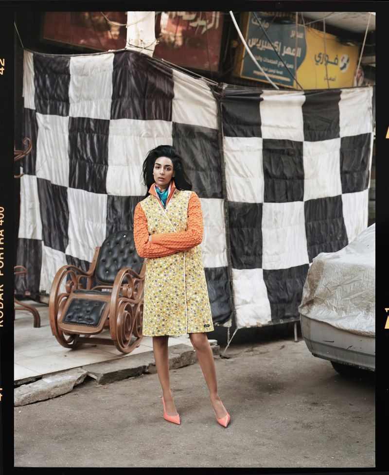Model Nora Attal stands outdoors wearing a yellow patterned dress over an orange knit sweater in front of a checkered backdrop.