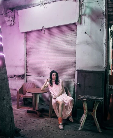 Model Nora Attal seated in a pale pink draped dress by outdoor tables in front of a closed street shutter under pink neon light.