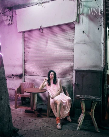 Model Nora Attal seated outdoors by a closed metal shutter wearing a draped pale pink dress and patterned mules in pink lighting.