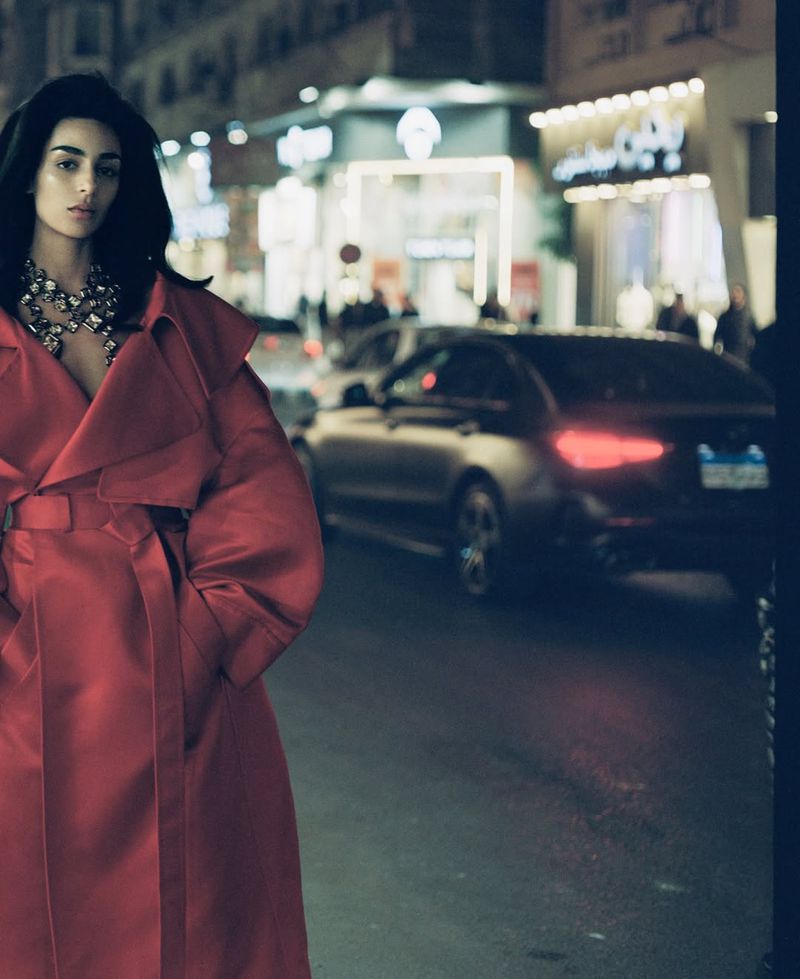 Model Nora Attal poses in a red belted trench coat on an urban street at night with city lights and a car in the background.