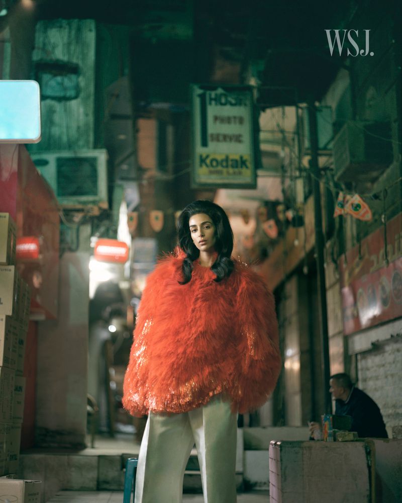 Model Nora Attal standing in an urban alley wearing an oversized orange textured coat and cream trousers for WSJ Magazine.