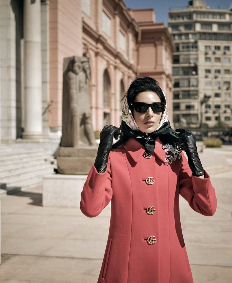 Model Nora Attal in a coral coat, black leather gloves, sunglasses, and a printed silk headscarf posing in downtown Cairo.