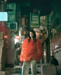 Model Nora Attal stands in an urban alleyway wearing a voluminous orange textured coat and cream wide-leg trousers.