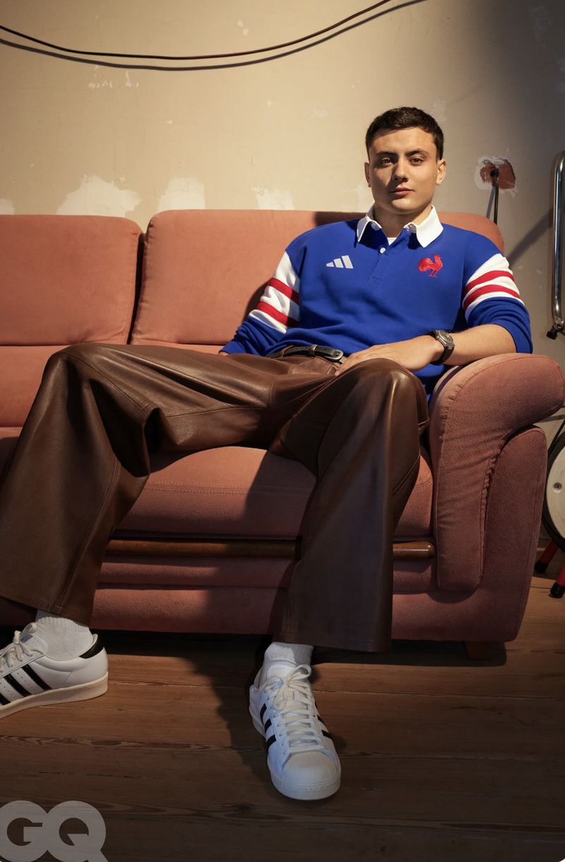 Louis Bielle-Biarrey seated on a pink couch wearing a blue rugby shirt, wide brown leather pants, and white sneakers.