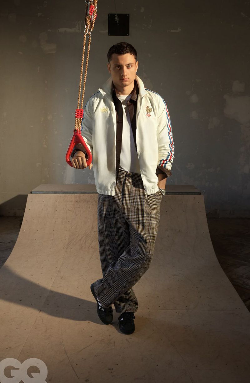 Louis Bielle-Biarrey standing on a wooden skate ramp holding a red gymnastic ring in a white track jacket and plaid trousers.