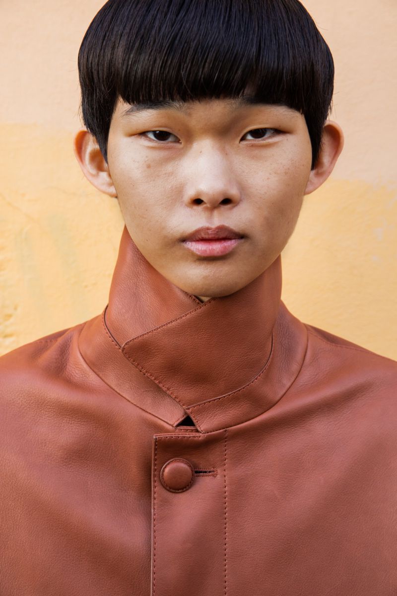 Close-up portrait of an East Asian male model with a bowl cut wearing a high-collar brown leather jacket against a warm yellow background.