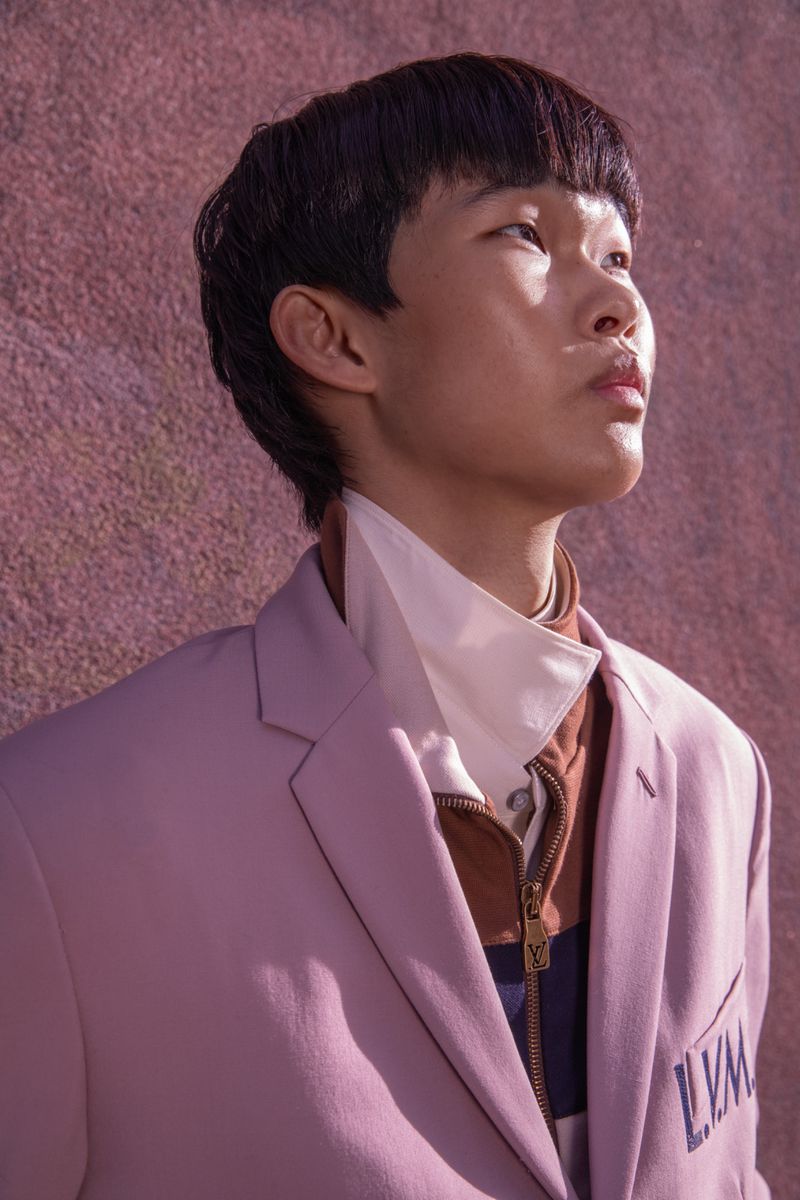 East Asian young man wearing a tailored lilac blazer and layered collar looking upward against a textured pink-mauve wall.