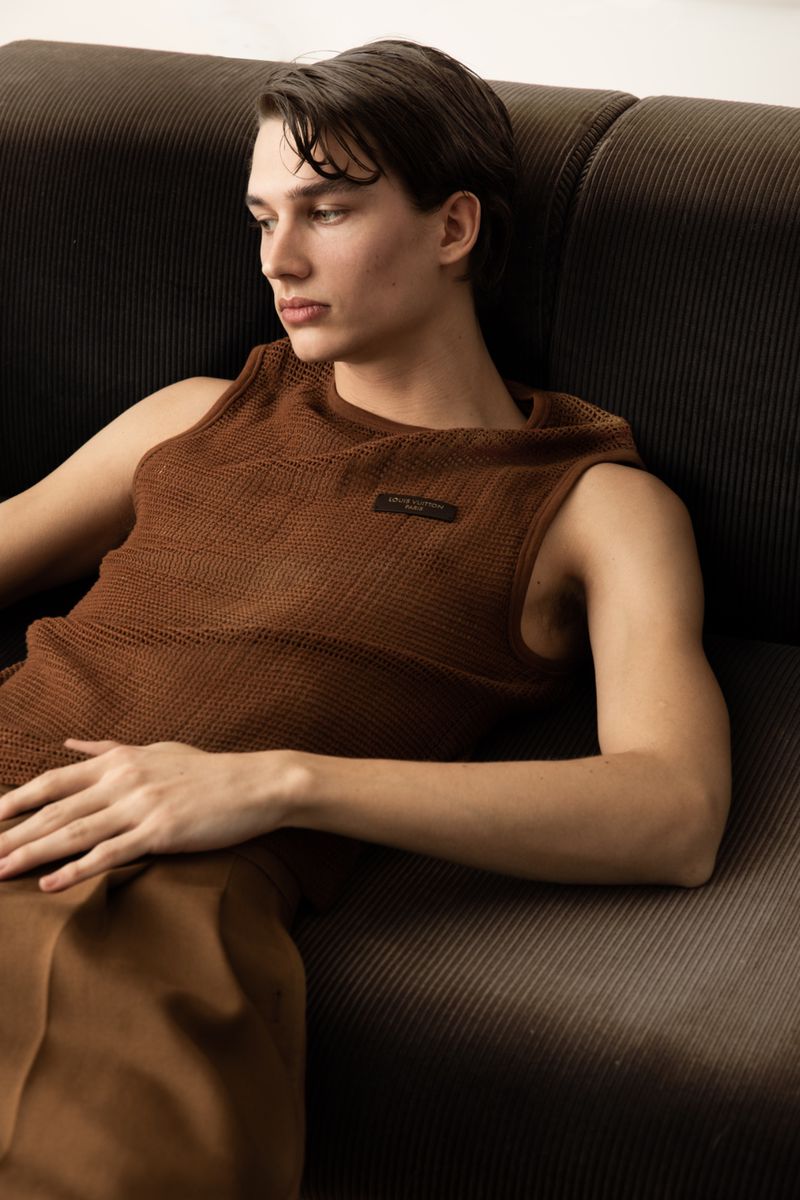 Young man in a brown sleeveless knit top reclining on a dark brown ribbed corduroy sofa.