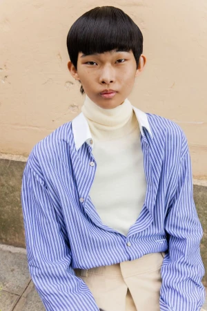 East Asian model with a bowl haircut wearing a blue striped button-down shirt over an off-white turtleneck sweater.