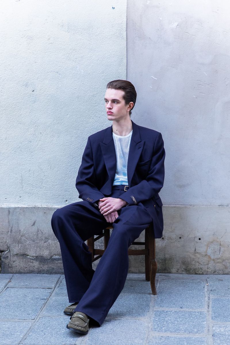A young man seated on a wooden chair wearing an oversized navy blue suit and loafers against a pale textured wall.