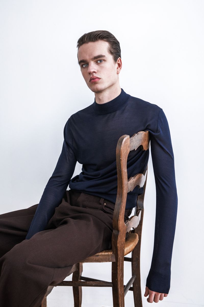Man in a navy mock-neck top with elongated sleeves and brown trousers seated backwards on a rustic wooden chair.
