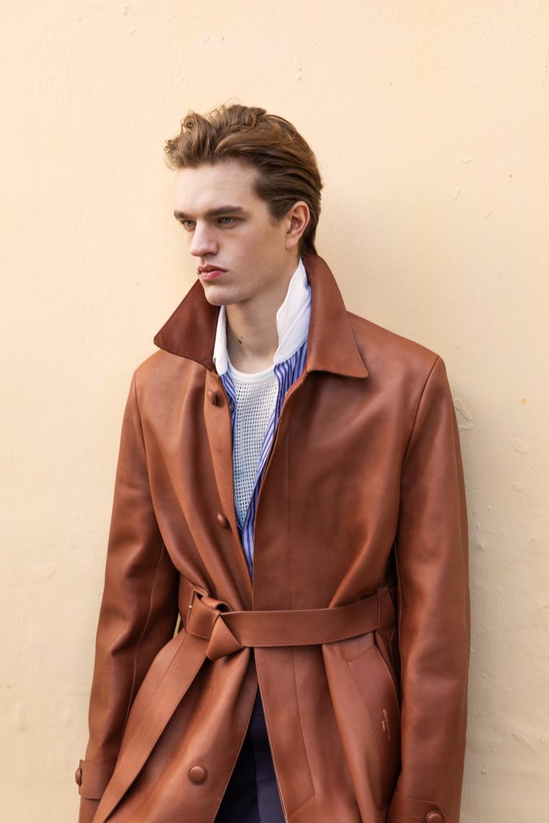 A male model wearing a belted brown leather coat over layered collared shirts against a warm beige background.