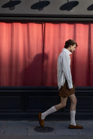 A man in an oversized white button-down shirt, brown shorts, white socks, and loafers walks in profile along a street facade.