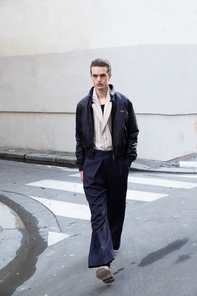 Fashion model walking on a paved street wearing a dark bomber jacket, wide-leg trousers, and a collared striped top.