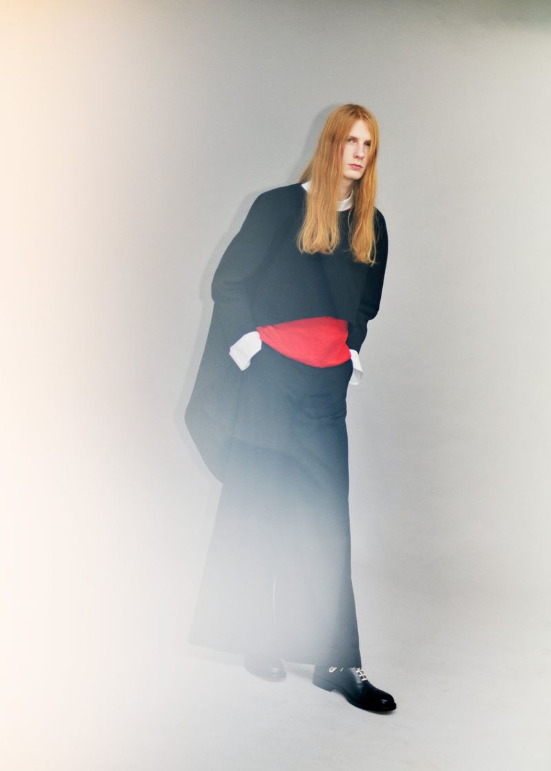 Model Filip Bryndza standing in layered black garments with a red waist accent against a neutral studio backdrop.