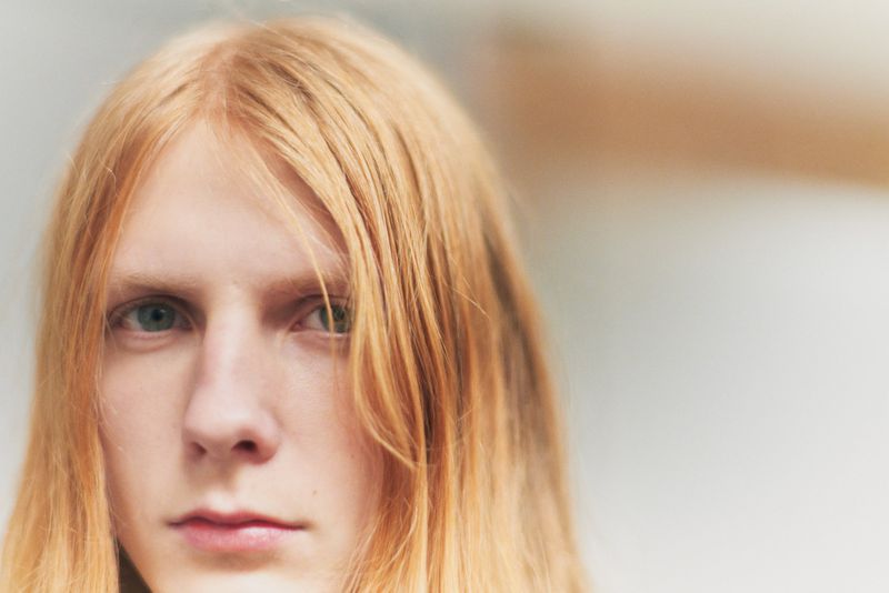 Close-up portrait of model Filip Bryndza with long reddish blonde hair looking directly forward against a soft neutral background.