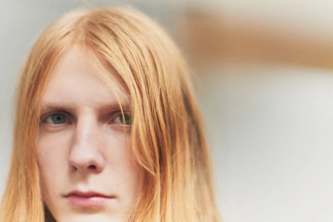 Close-up portrait of model Filip Bryndza with long reddish blonde hair looking directly forward against a soft neutral background.