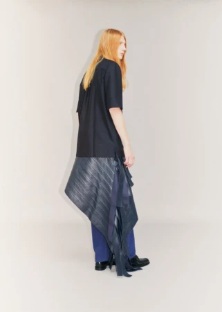 Model Filip Bryndza wearing a black short-sleeved shirt, asymmetrical pleated wrap, and blue trousers against an off-white background.