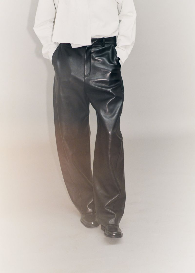 Cropped view of a model wearing wide-leg black leather trousers and a loose white shirt against a neutral studio backdrop.