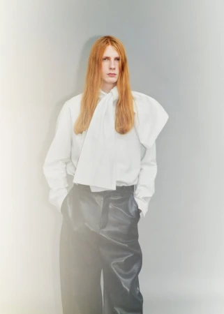 Model Filip Bryndza wearing an oversized white pussy-bow blouse and black leather trousers against a grey studio backdrop.