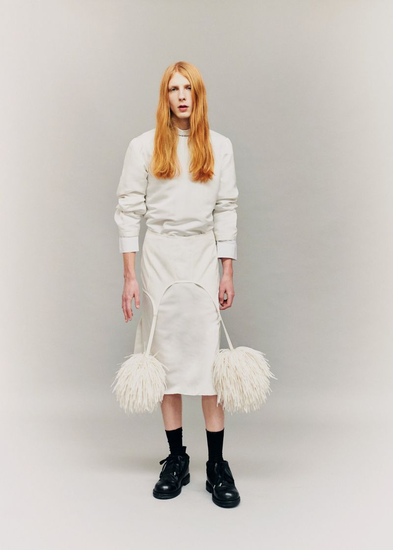 Filip Bryndza standing in an all-white skirt ensemble with feather details and black boots against a grey studio backdrop.