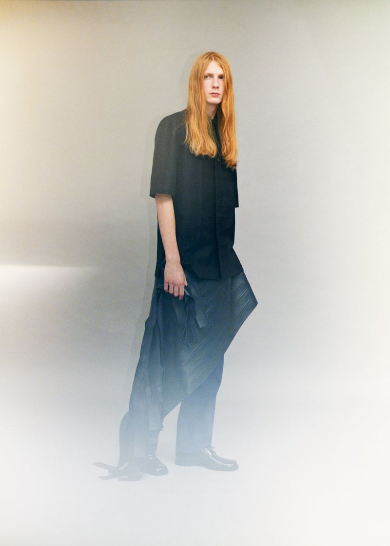 Model Filip Bryndza with long strawberry blonde hair wearing an all-black shirt and pleated wrap trousers in a minimal studio setting.