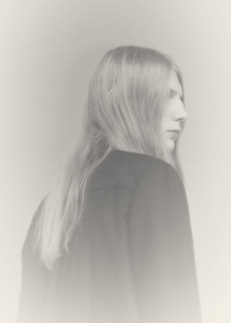 Model Filip Bryndza with long blonde hair viewed from behind in a profile angle wearing a dark jacket against a soft pale backdrop.