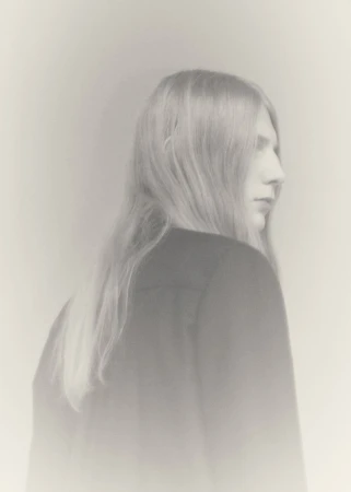 Model Filip Bryndza with long blonde hair viewed from behind in a profile angle wearing a dark jacket against a soft pale backdrop.