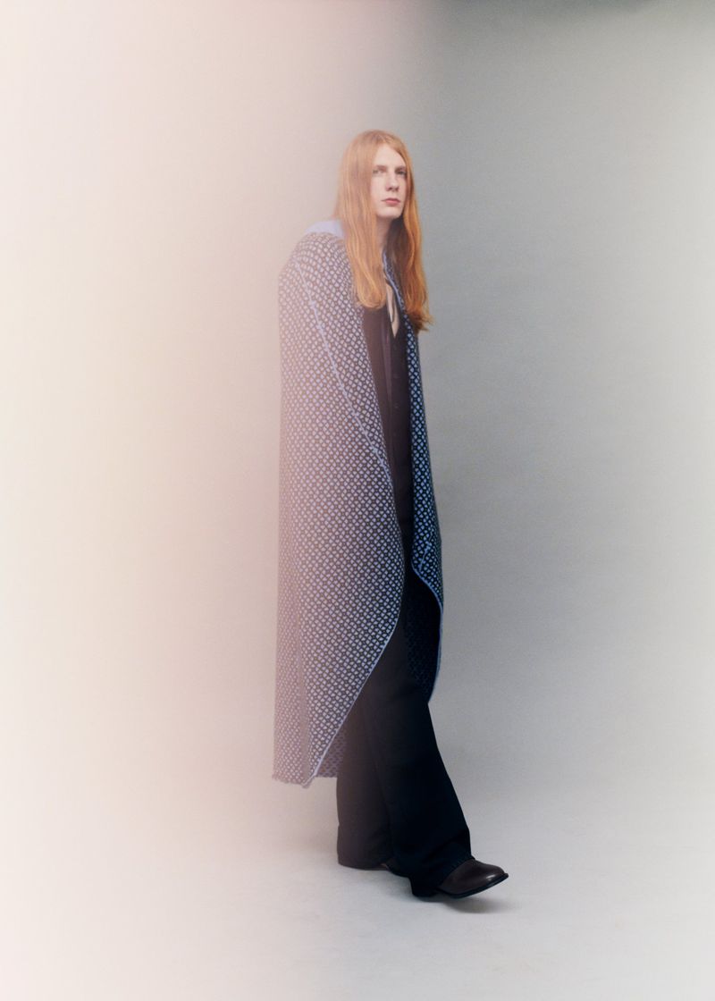 Model Filip Bryndza wearing a long patterned draped cape and wide-leg dark trousers against a neutral studio backdrop.