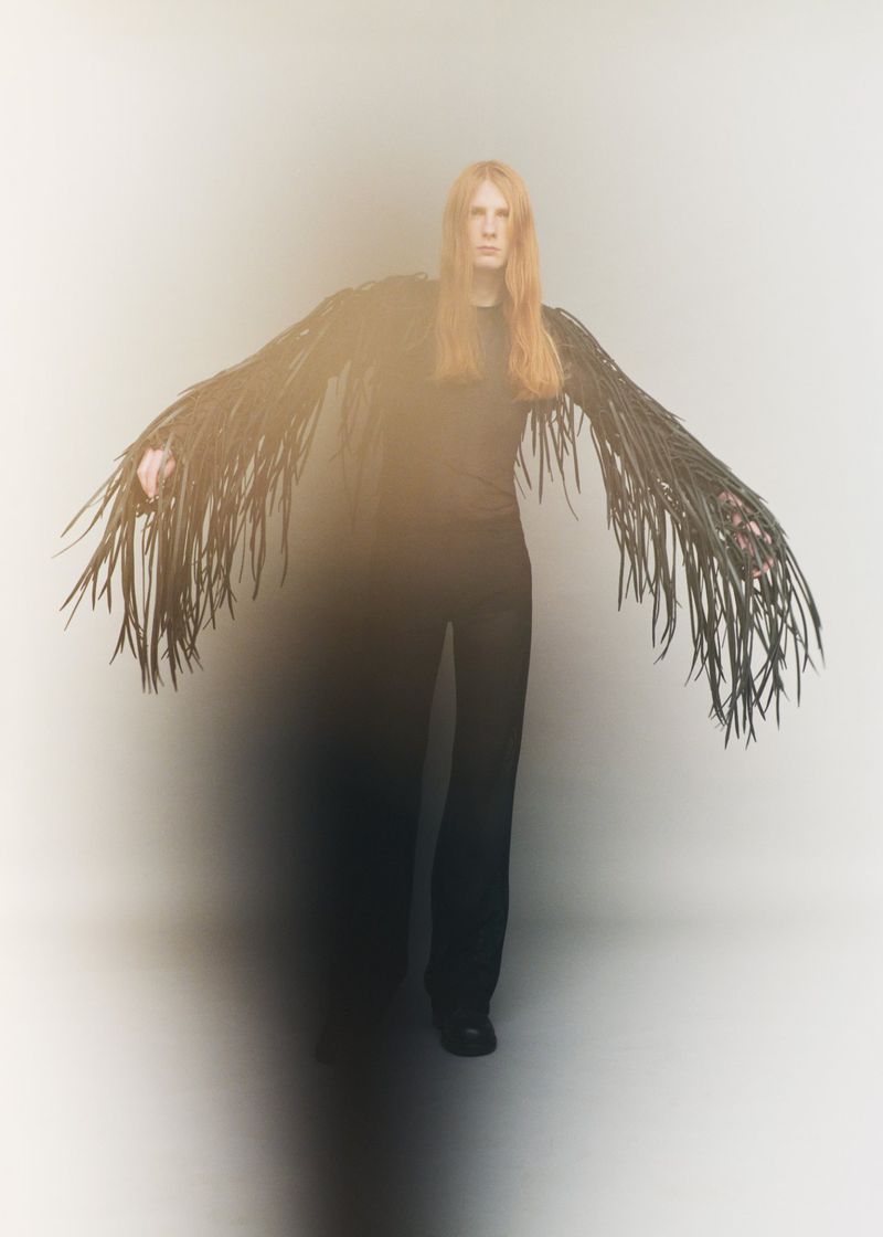 Model with long reddish hair wearing an all-black outfit with dramatic shredded fringe sleeves against a soft grey studio backdrop.