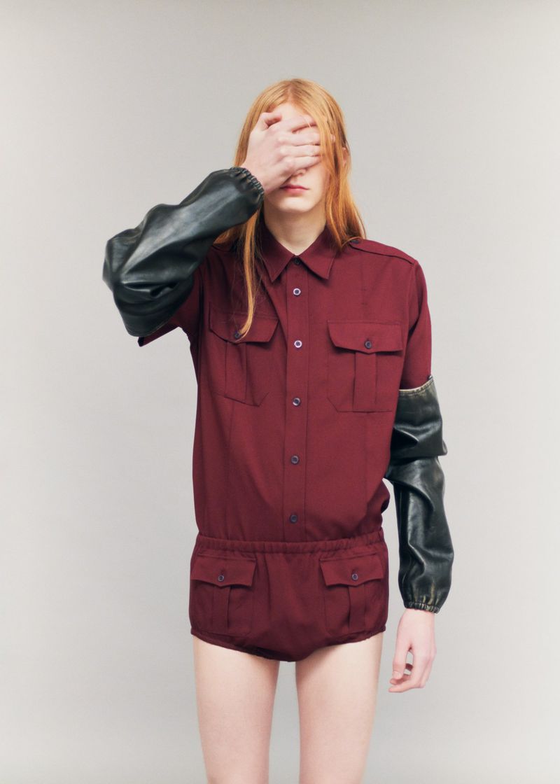 Model with long reddish-blonde hair covering face with hand, wearing a burgundy utility shirt and matching shorts.