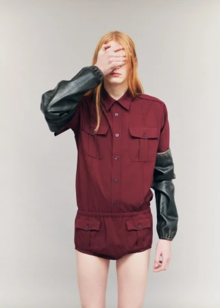 Model with long reddish-blonde hair covering face with hand, wearing a burgundy utility shirt and matching shorts.