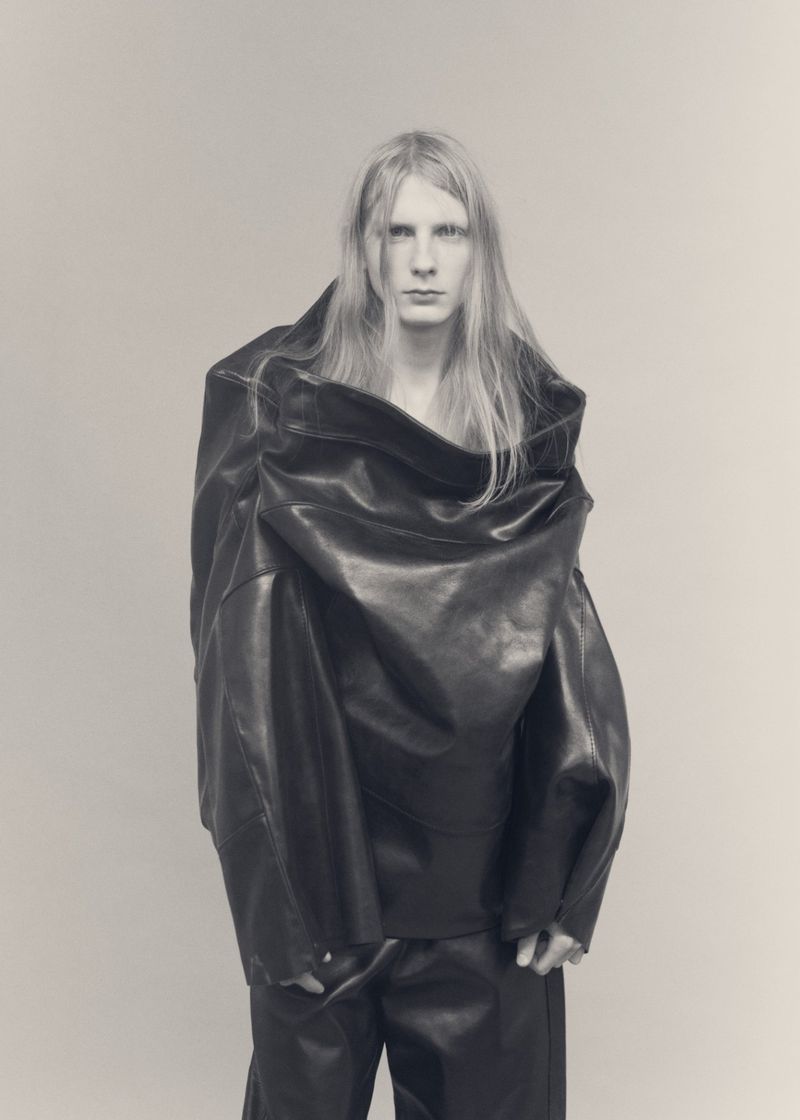 Model Filip Bryndza wearing an oversized draped black leather garment against a plain grey studio backdrop.