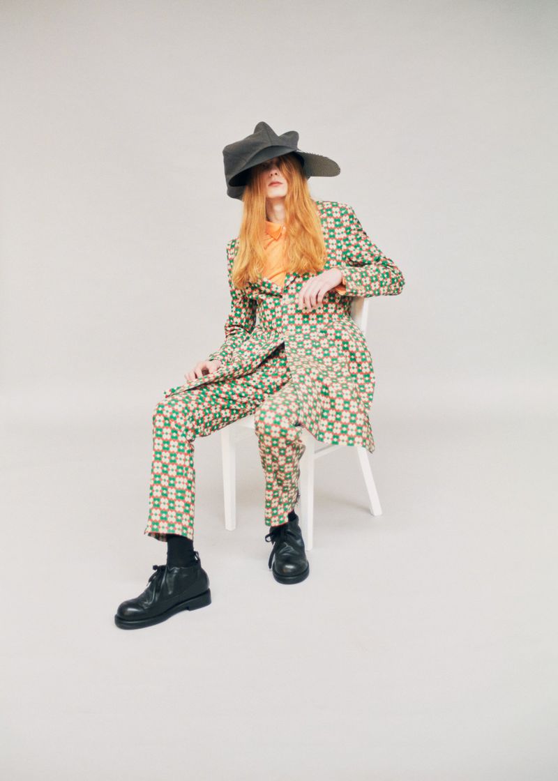Model Filip Bryndza seated on a white chair wearing a green patterned suit and oversized dark hat against a studio backdrop.